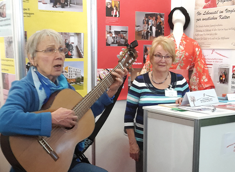 eine Wohnfrau spielt am Infostand Gitarre, die andere hört zu