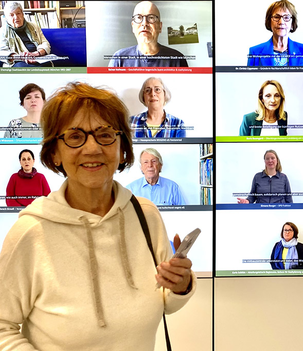 Christa Lippmann beim 10 jährigem Jubiläum der Mitbauzentrale, vor einer Videowand mit Abbildungen von Personen, die über Ihre Erfahrungen sprechen